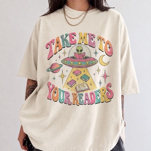 May include: A beige short-sleeve t-shirt featuring a graphic of an alien in a flying saucer reading a book. The text "TAKE ME TO YOUR READERS" is printed in colorful, wavy letters. The design includes planets, stars, and floating books.
