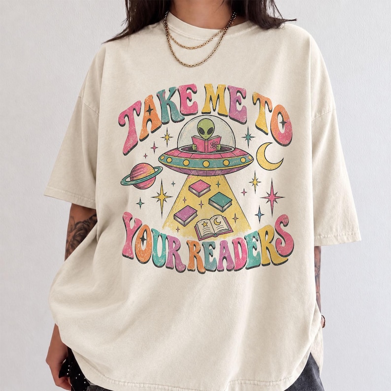 May include: A beige short-sleeve t-shirt featuring a graphic of an alien in a flying saucer reading a book. The text "TAKE ME TO YOUR READERS" is printed in colorful, wavy letters. The design includes planets, stars, and floating books.