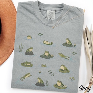 May include: A grey short-sleeve t-shirt featuring a Comfort Colors label at the neck and a graphic print of various green frogs in different poses, including swimming, sitting on lily pads, and resting on rocks, surrounded by small water plants.