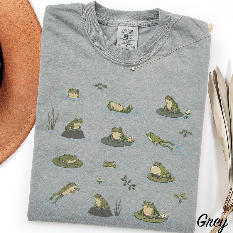 May include: A grey short-sleeve t-shirt featuring a Comfort Colors label at the neck and a graphic print of various green frogs in different poses, including swimming, sitting on lily pads, and resting on rocks, surrounded by small water plants.