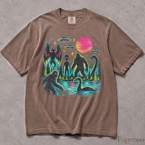 May include: A brown short-sleeve t-shirt featuring a colorful graphic print of a mothman, an alien, a bigfoot, and a loch ness monster near a lake with pine trees, UFOs, and a pink sun. The shirt has a crew neckline and a Comfort Colors brand label.