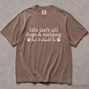 May include: A brown short-sleeve t-shirt with white text that reads, "life isn't all dogs & mahjong but it should be," accompanied by two white paw print graphics. The shirt features a crew neckline and a visible brand tag at the inner collar.