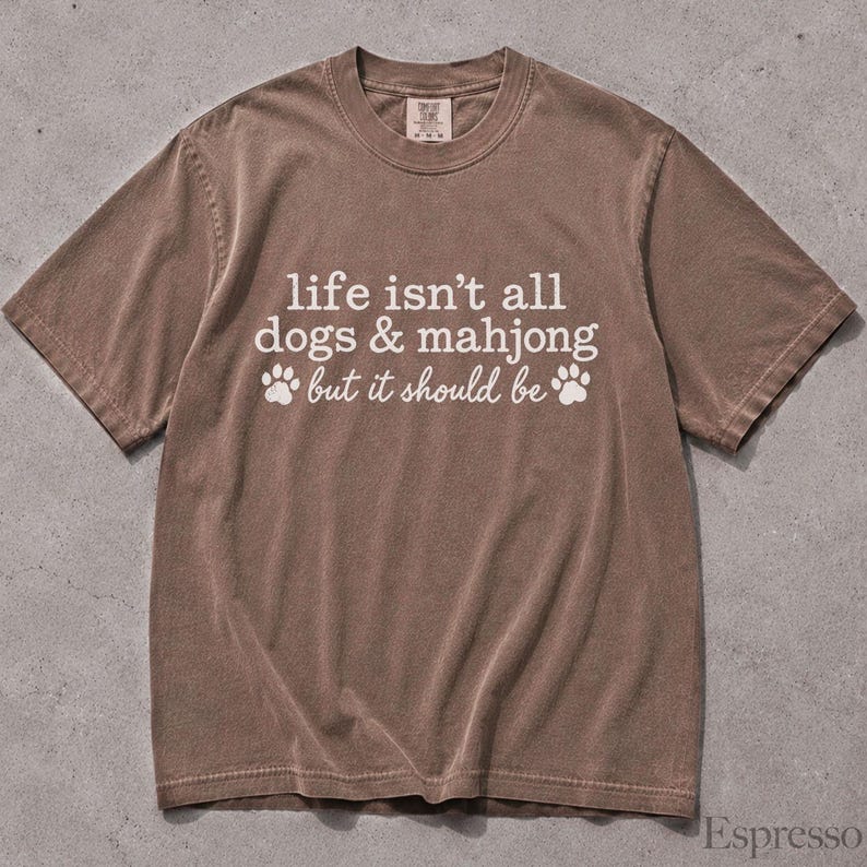 May include: A brown short-sleeve t-shirt with white text that reads, "life isn't all dogs & mahjong but it should be," accompanied by two white paw print graphics. The shirt features a crew neckline and a visible brand tag at the inner collar.