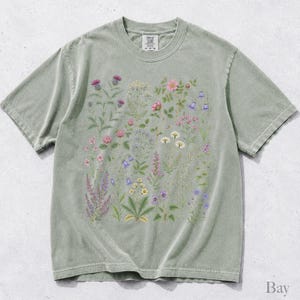 May include: A sage green short-sleeve t-shirt featuring a central graphic print of various wildflowers and botanical illustrations in shades of pink, purple, yellow, and white. The shirt has a crew neckline and a visible brand label at the inner collar.