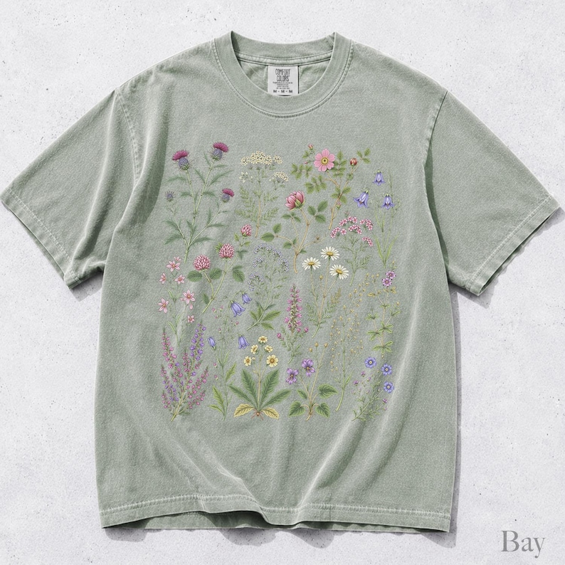 May include: A sage green short-sleeve t-shirt featuring a central graphic print of various wildflowers and botanical illustrations in shades of pink, purple, yellow, and white. The shirt has a crew neckline and a visible brand label at the inner collar.