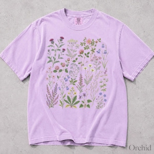 May include: A light purple short-sleeve t-shirt featuring a central graphic print of various wildflowers and botanical illustrations in shades of pink, blue, yellow, and green. The shirt has a crew neckline and a label at the inner neck area.
