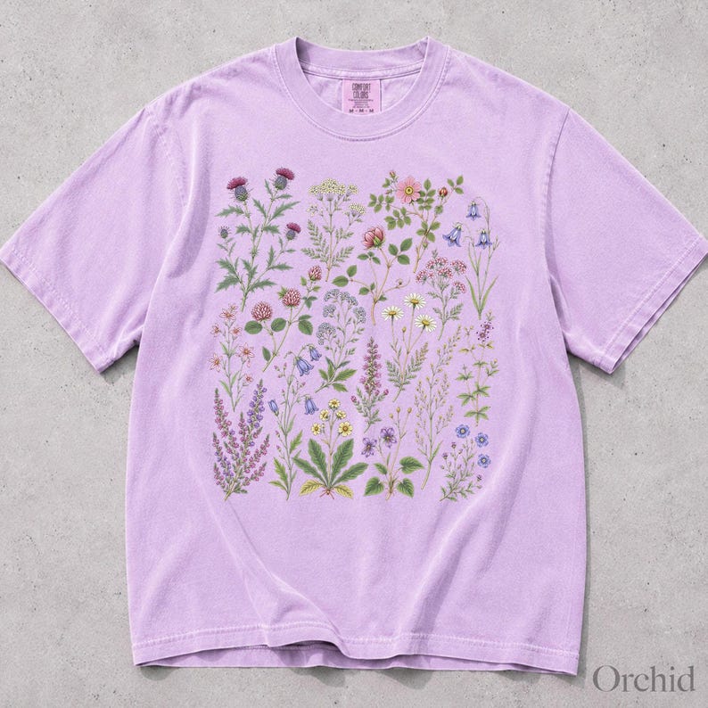 May include: A light purple short-sleeve t-shirt featuring a central graphic print of various wildflowers and botanical illustrations in shades of pink, blue, yellow, and green. The shirt has a crew neckline and a label at the inner neck area.