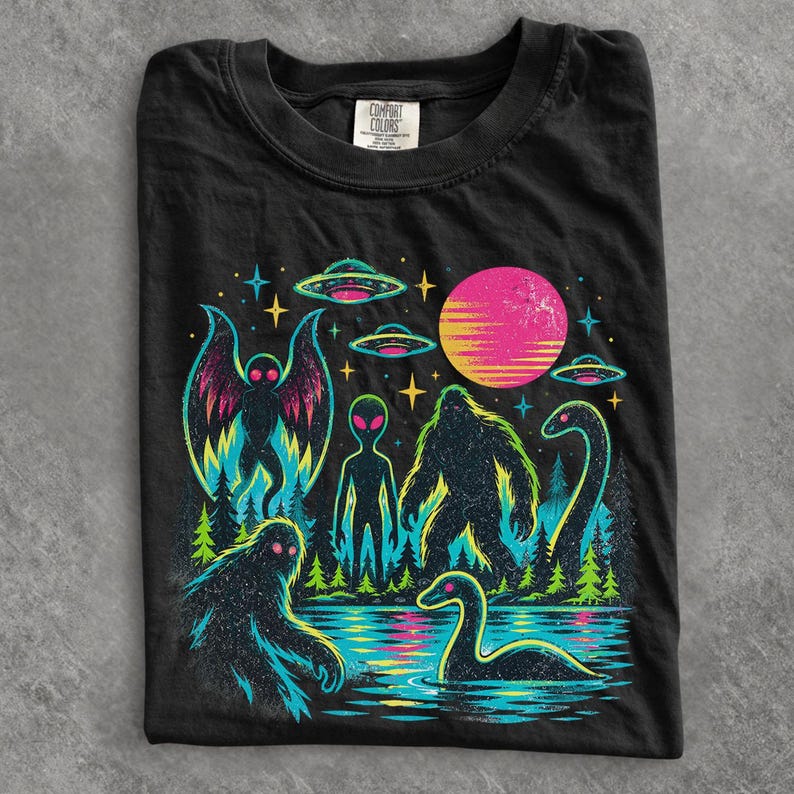 May include: A black t-shirt featuring a vibrant graphic print of cryptids and extraterrestrials, including a moth-like creature, an alien, a sasquatch, and a sea serpent, set against a forest landscape with UFOs, stars, and a large pink sun.