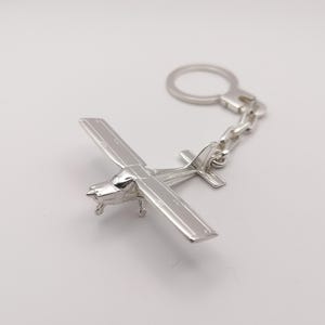 Op de afbeelding: Zilveren vliegtuig sleutelhanger met een gedetailleerd ontwerp. De sleutelhanger heeft een zilveren ketting en een zilveren ring.