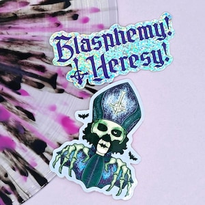 May include: Two stickers on a pink background. One sticker has the text "Blasphemy! Heresy!" in purple glitter. The other sticker is a skull wearing a bishop's hat with bats.