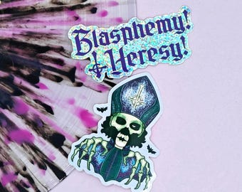 May include: Two stickers on a pink background. One sticker has the text "Blasphemy! Heresy!" in purple glitter. The other sticker is a skull wearing a bishop's hat with bats.