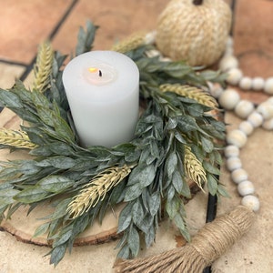 May include: A white candle sits in the centre of a green and beige wreath on a wooden slice. The wreath is surrounded by white beads and a brown jute tassel.