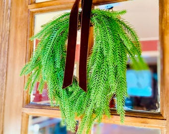 May include: A vibrant green wreath with long, needle-like leaves hangs from a dark burgundy velvet ribbon. The wreath is displayed on a wooden-framed glass door, creating a welcoming and festive look.