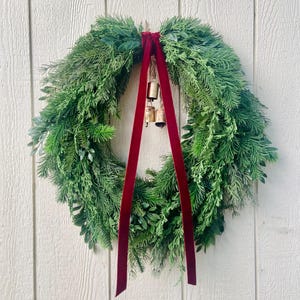 Könnte beinhalten: Ein runder grüner Kranz mit einem tiefroten Samtband und kleinen goldenen Glocken. Der Kranz besteht aus verschiedenen grünen Blättern und hängt vor einem weißen Holzgrund. Eine festliche Weihnachtsdekoration.