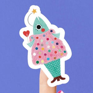 May include: A quirky sticker of a fish character with a teal head, a pink dress with colourful polka dots, and brown boots. The fish holds a heart-shaped lollipop and has a star above its head. The sticker is set against a blue backdrop.