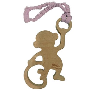May include: Wooden baby toy in the shape of a monkey with a light pink fabric loop. The monkey has a cutout tail and is holding a ring. The text "gugu the hero" is printed on the monkey.