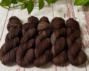 May include: A collection of brown yarn skeins with varying shades of dark brown, light brown, and hints of purple. The yarn is arranged on a light-colored wooden surface, with green leaves in the background. The yarn appears soft and textured.