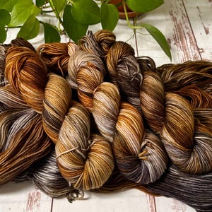 May include: A collection of hand-dyed yarn skeins in shades of brown, tan, and gray. The yarn is arranged on a light-colored wooden surface, with a green plant in the background. The yarn has a variegated color pattern, creating a textured appearance.