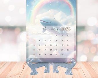 May include: A blue whale with a rainbow above it is featured on a January 2025 calendar. The calendar is on a blue metal stand.