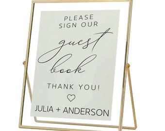 May include: A rectangular sign in a thin gold metal frame with a stand. The white sign features black text that reads: PLEASE SIGN OUR guest book THANK YOU! followed by a heart icon and the names JULIA + ANDERSON in a clean, modern font.