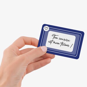 May include: A rectangular card with a dark blue border and a white interior, held by a hand. The card features the text "Ton sourire est mon tr&eacute;sor !" in elegant script, with a small heart icon in the upper left corner.