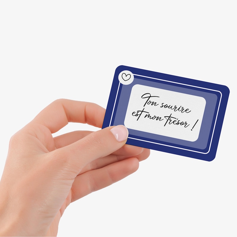 May include: A rectangular card with a dark blue border and a white interior, held by a hand. The card features the text "Ton sourire est mon tr&eacute;sor !" in elegant script, with a small heart icon in the upper left corner.