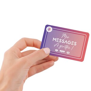 May include: A rectangular card with a gradient purple to pink background is held in a hand. The card has white text that reads "Mes MESSAGES A gratter!" with icons below. The card has a white border and a circular logo in the upper left corner.