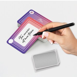 May include: A stack of purple and pink cards with the text "Ton sourire est mon tr&eacute;sor" in black script. A black pen is held over the card. A small stack of silver cards is in the foreground.