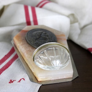 Puede incluir: Un pequeño soporte rectangular de mármol rosa y blanco con una base de metal plateado. El soporte tiene un pequeño plato de vidrio transparente con un borde dorado y un medallón de metal con una imagen en relieve de una cara.