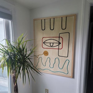 May include: A framed abstract art piece with a wooden frame. The artwork features a stylised eye with a red outline, a yellow circle, and blue wavy lines on a beige background.