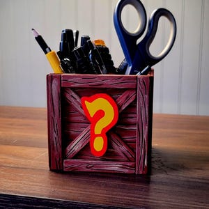 May include: A wooden pencil holder shaped like a crate with a question mark on the front. The crate is painted brown with a red stain. The question mark is yellow with a red outline. The holder is filled with pens, pencils, and a pair of scissors.