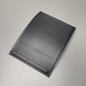 Könnte beinhalten: Ein rechteckiges, dunkelgraues Objekt mit geriffelter Oberfläche. Die parallelen Rillen erzeugen ein strukturiertes Muster. Die Kanten weisen kleine, gleichmäßig verteilte Vorsprünge auf. Das Objekt scheint aus einem festen Material zu bestehen.