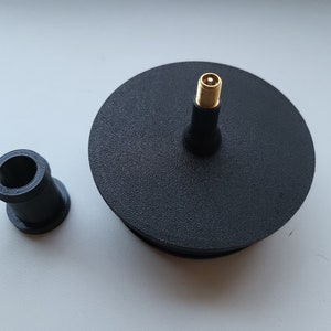 Könnte beinhalten: Eine schwarze, kreisförmige Komponente mit einem goldfarbenen Stecker und einem separaten schwarzen zylindrischen Teil. Das kreisförmige Teil hat eine strukturierte Oberfläche. Die Teile sind wahrscheinlich elektronische Komponenten oder Teile.