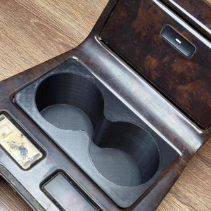 Op de afbeelding: Een houten interieurpaneel voor een auto met twee bekerhouders en een knop voor de gevarendriehoek. Het paneel heeft een houtnerf afwerking en een zwarte kunststof rand.
