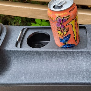 Op de afbeelding: Een zwarte bekerhouder voor in de auto met een blikje Monster Energy-drank erin. Het blikje is oranje en heeft een vlinderontwerp met de tekst "Monster Energy" en "Taurine" erop.