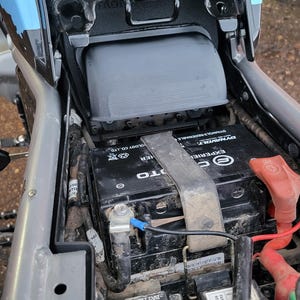 Op de afbeelding: Een motoraccucompartiment met een zwarte Dynavolt-accu vastgezet met een metalen band. De assemblage bevat een rode pluspoolafdekking, een zwarte minkabel en twee kleine zwarte relaisboxen met het label Fan Relay en Spark Relay.