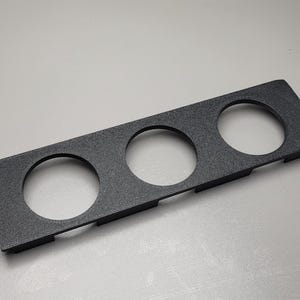 Può includere: Un pannello rettangolare nero con finitura testurizzata dotato di tre fori circolari equidistanti. Il pannello presenta una superficie superiore piatta e bordi laterali corti e perpendicolari che si estendono verso il basso.