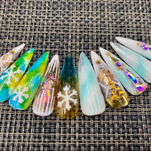 Puede incluir: Un conjunto de diez uñas postizas largas y puntiagudas con varios diseños de temática invernal. Los diseños incluyen copos de nieve, purpurina y un degradado de azul y blanco.
