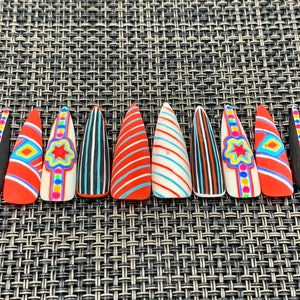 May include: A set of ten long, pointed false nails. The nails feature various designs, including red and white stripes, black and white stripes, and geometric patterns with bright colors like blue, yellow, and orange. The nails are arranged on a textured surface.