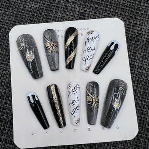 May include: A collection of ten false nails with diverse designs. The nails display glitter, bows, and the text "Happy new year." Colours include black, white, and silver. Some nails feature a champagne glass design.