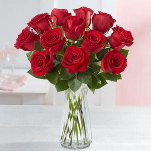 Puede incluir: Un ramo de una docena de rosas rojas dispuestas en un jarrón de vidrio transparente. Las rosas están en plena floración y tienen una forma clásica.