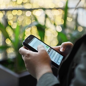 May include: A person holding a black smartphone with a touchscreen display. The screen shows a text message interface with a virtual keyboard. The background is blurred with bokeh lights.