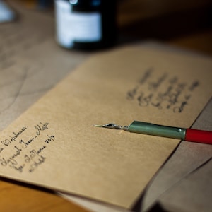 May include: A brown envelope with handwritten text on it, a dip pen with a red and green handle, and a glass ink bottle.