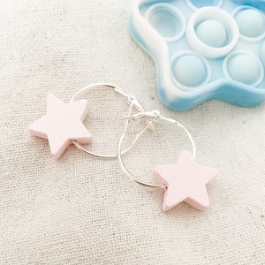 Può includere: Un paio di orecchini a cerchio d'argento con ciondoli a forma di stella rosa. Gli orecchini sono su uno sfondo di tessuto bianco.