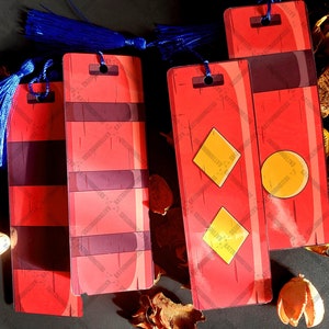 Könnte beinhalten: Vier rot-schwarze gestreifte Lesezeichen mit blauen Quasten. Die Lesezeichen haben verschiedene Designs, darunter einen gelben Kreis und zwei gelbe Diamanten.