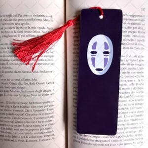 Könnte beinhalten: Ein schwarzes Lesezeichen mit einem weißen Quasten. Das Lesezeichen zeigt ein weißes Gesicht mit schwarzem Umriss und zwei blauen Augen. Das Gesicht stammt aus dem Animationsfilm Chihiros Reise ins Zauberland.