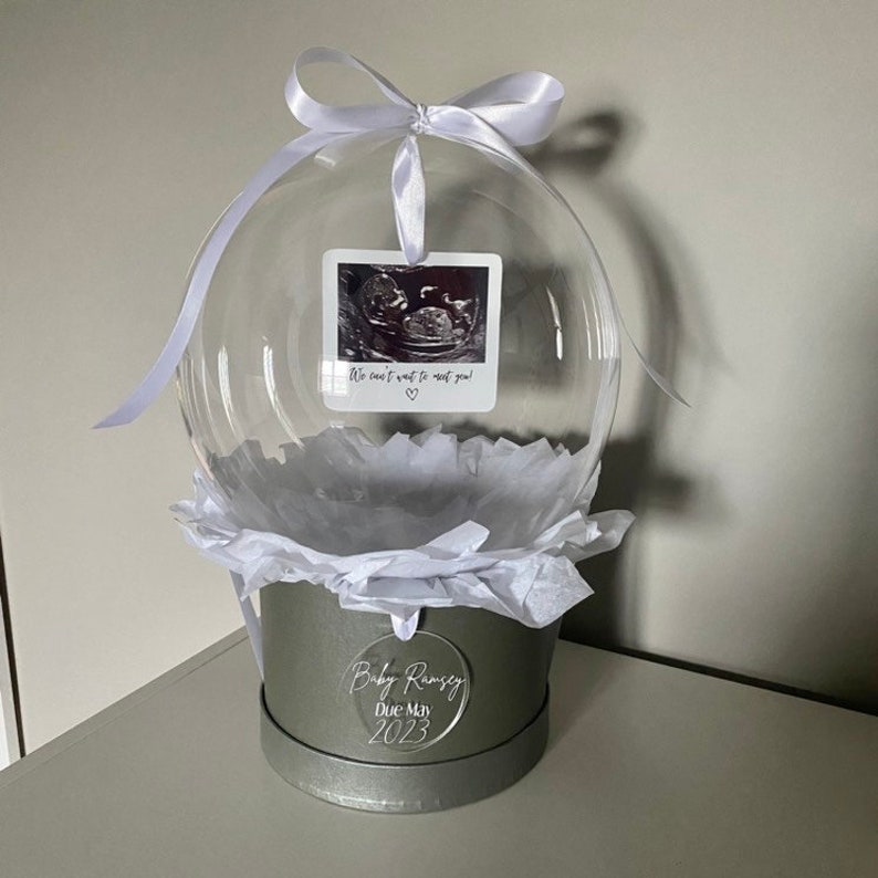 May include: A clear glass globe with a white ribbon and a black and white ultrasound image inside. The globe is sitting on a gray base with a white circle that says "Baby Ramsey Due May 2023".