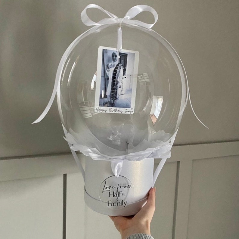 May include: A white balloon with a clear plastic bubble containing a photo and the text "Happy Birthday Imaja". The balloon is attached to a white box with a white ribbon and a white tag that says "Love from Haifa & Family".