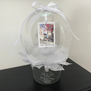 May include: A clear balloon with a white ribbon and a photo card inside. The card has a photo of a person and the text "Happy 30th Birthday!" The balloon is sitting on a white tissue paper base and a gray box with the text "Lots of love - Mum, Dad, Asha, Robin & Harsh".