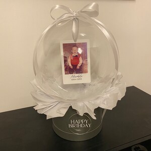 May include: A clear balloon with a white ribbon and a photo inside. The photo shows a young boy wearing a red shirt and a bow tie. The balloon is sitting on a white tissue paper base with a black and white label that says "Happy Birthday".